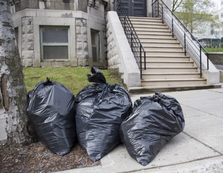 Three large black plastic rubbish bags filled with waste are placed on the pavement near the base of a tree, adjacent to a concrete sidewalk outside a building with a staircase. The bags are tightly knotted at the tops and appear full, with some bulging and irregular shapes indicating contents inside. The surrounding environment includes a small patch of grass and a tree trunk to the left of the bags, with the building in the background having a rounded stone exterior and a few small windows. A broad concrete staircase with black metal handrails leads up from the sidewalk to an entrance and is situated directly behind the rubbish bags. The scene is captured in natural daylight, showing no other objects or vehicles nearby. The image reflects typical outdoor rubbish disposal near an urban residential area, aligning with rubbish removal services such as those provided by Commercial Waste Removal Palmers Green in London.