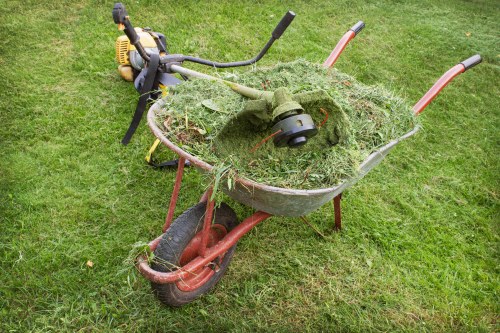 A red garden wheelbarrow filled with freshly cut grass and plant debris, positioned on a green lawn. The wheelbarrow has a black rubber wheel and metal handles. Resting on top of the grass in the wheelbarrow is a green petrol-powered grass trimmer with a black handle and an orange safety guard. The surrounding area consists of well-maintained grass, indicating an outdoor garden or backyard setting, with natural daylight illuminating the scene. The image is related to garden waste disposal services provided by Commercial Waste Removal Palmers Green, serving local residents and businesses in the area, including postal code N13.