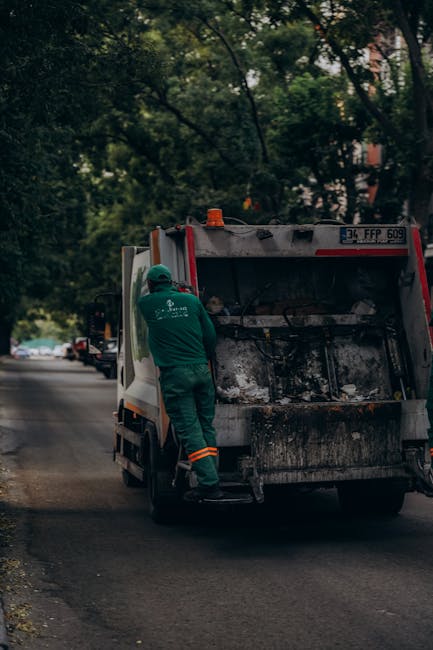 A street scene featuring a waste collection vehicle parked on the side of a road with a worker in green uniform and safety helmet actively engaged in the process of rubbish removal. The vehicle is a m