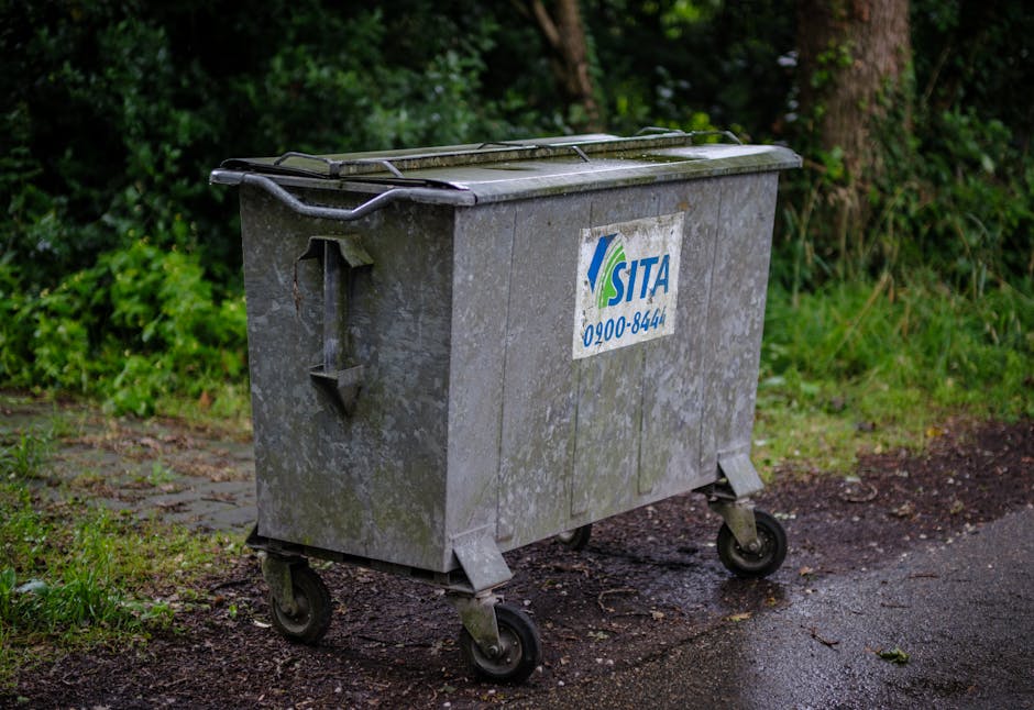 A large, rectangular metal wheelie bin with a slightly weathered, grey finish sits on a paved path adjacent to a grassy area with lush green bushes and trees in the background. The bin's surface shows