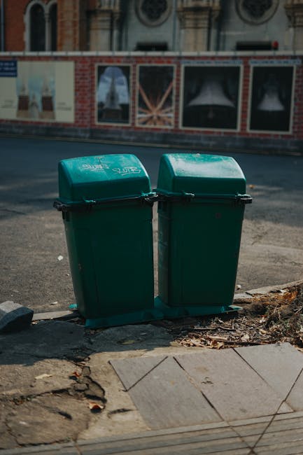 Two green outdoor rubbish bins with hinged lids, positioned side by side on a sidewalk curb. The bins are made of durable plastic with a smooth, slightly glossy finish and black locking mechanisms alo