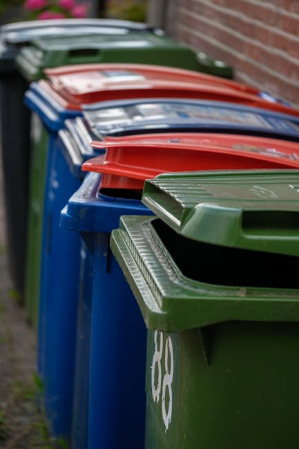 Two green outdoor rubbish bins with hinged lids, positioned side by side on a sidewalk curb. The bins are made of durable plastic with a smooth, slightly glossy finish and black locking mechanisms alo